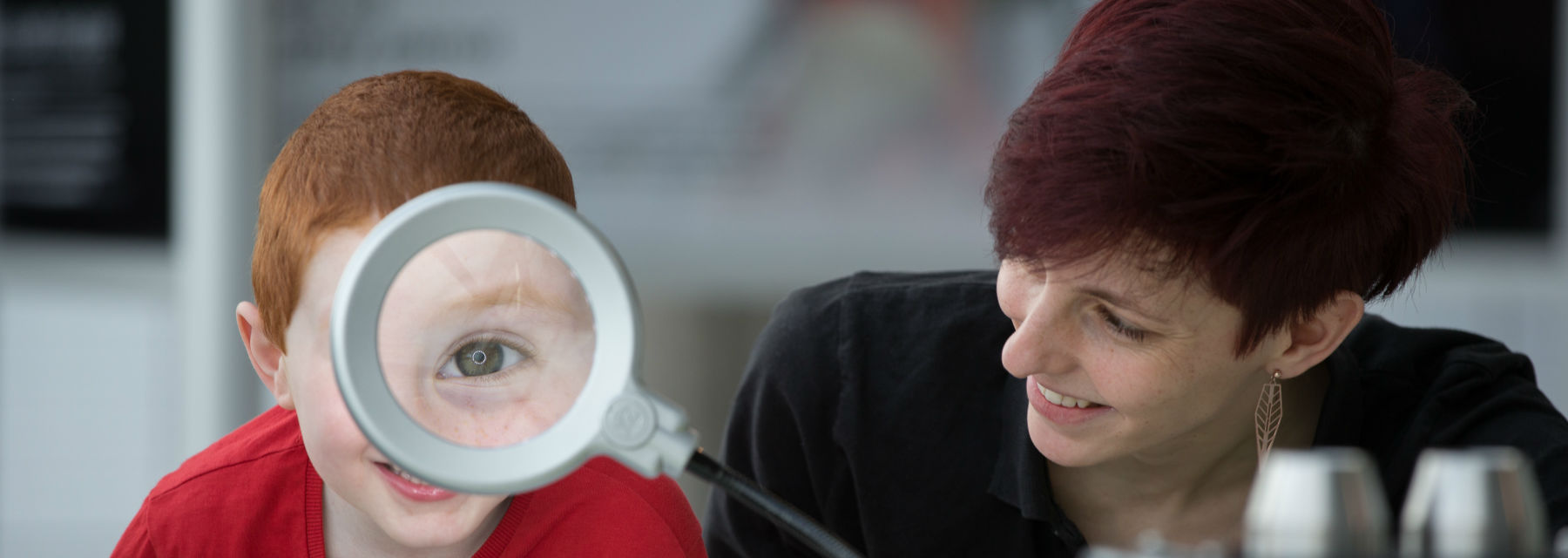 A boy's eye appears huge through a magnifying glass. An expert sits beside him.