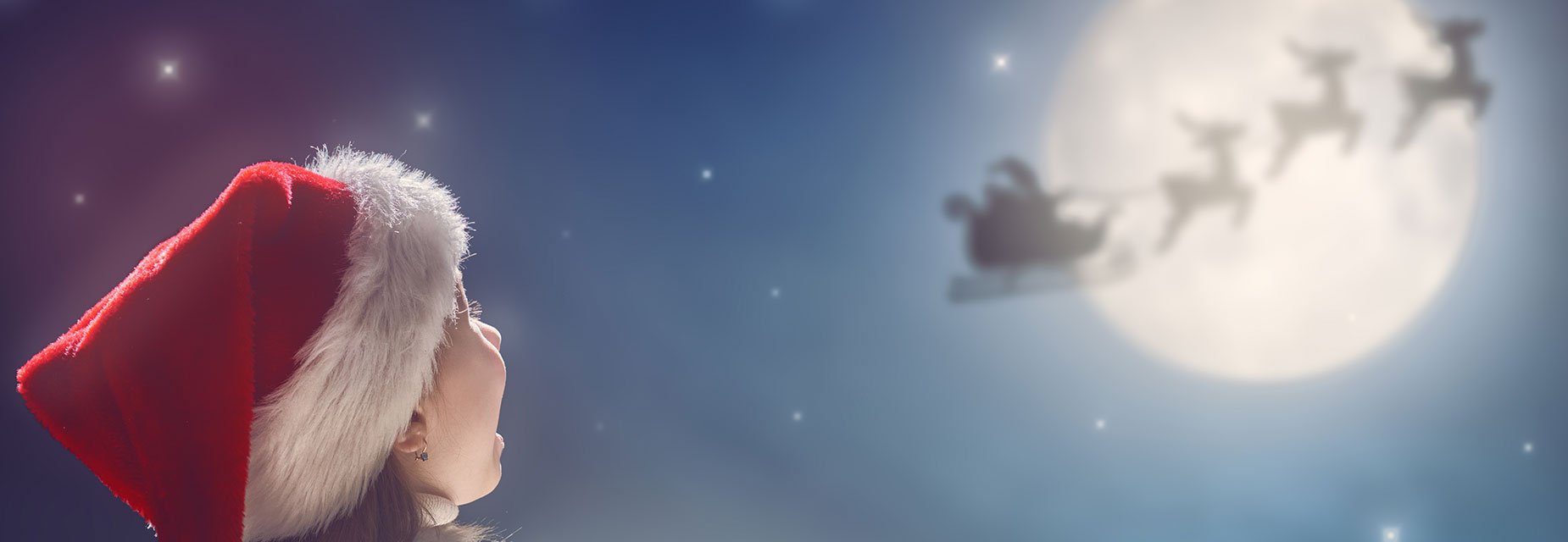 A young girl looks into the sky at a silhouette of Santa's sleigh against the moon