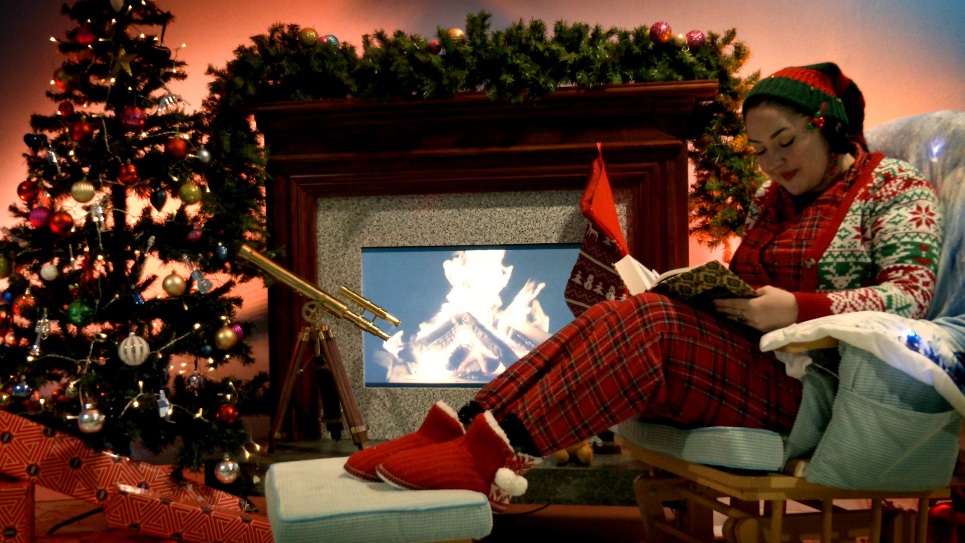 Veronica reads a book from a chair in front of a fire surrounded by Christmas decorations