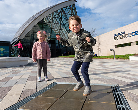 A child jumps up from a tiled square in front of the main entrance to Glasgow Science Centre. Image: Paul Upward and Timberplay Scotland 