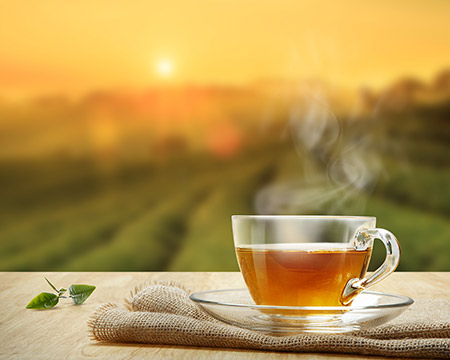 A cup of tea and tea leaf on an outside table set against the backdrop of the sun rising over a hillside tea plantation