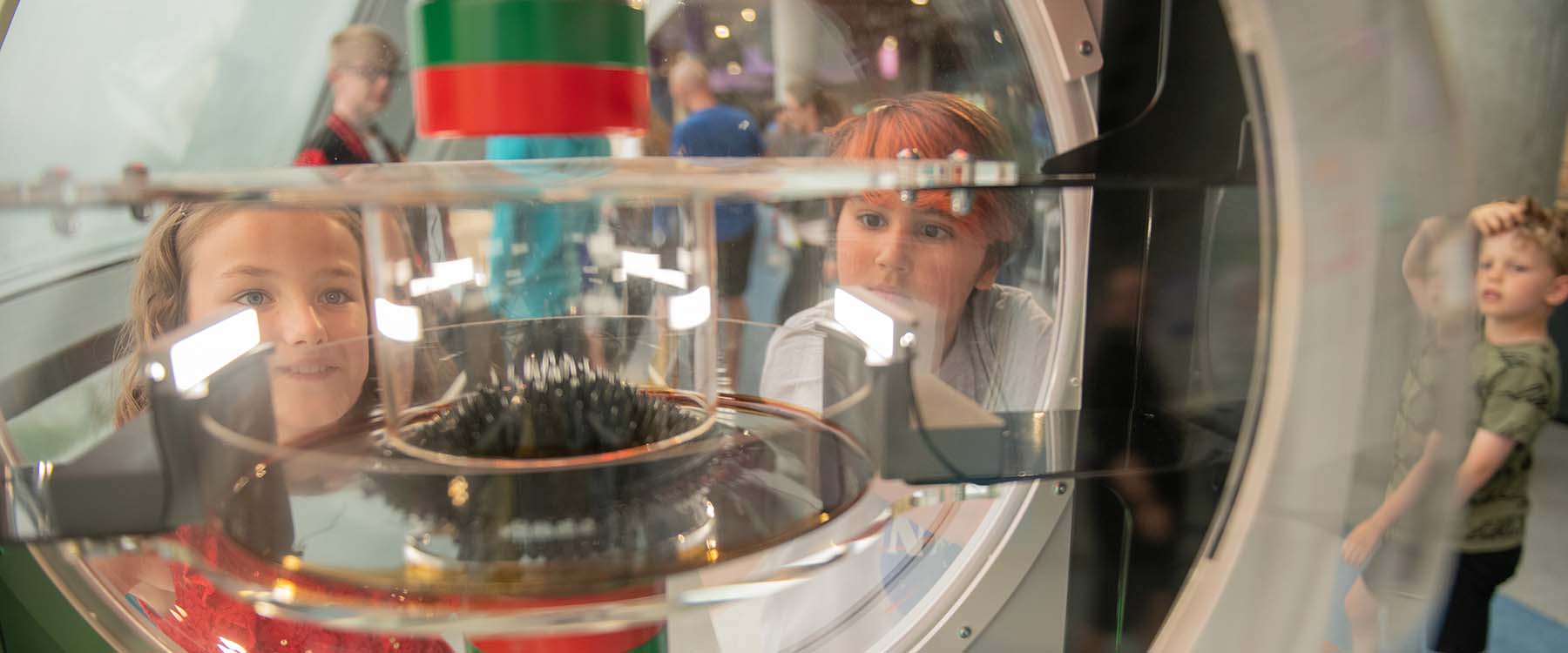 Young people look through to the shape of a magnetic liquid being distorted within transparent dome