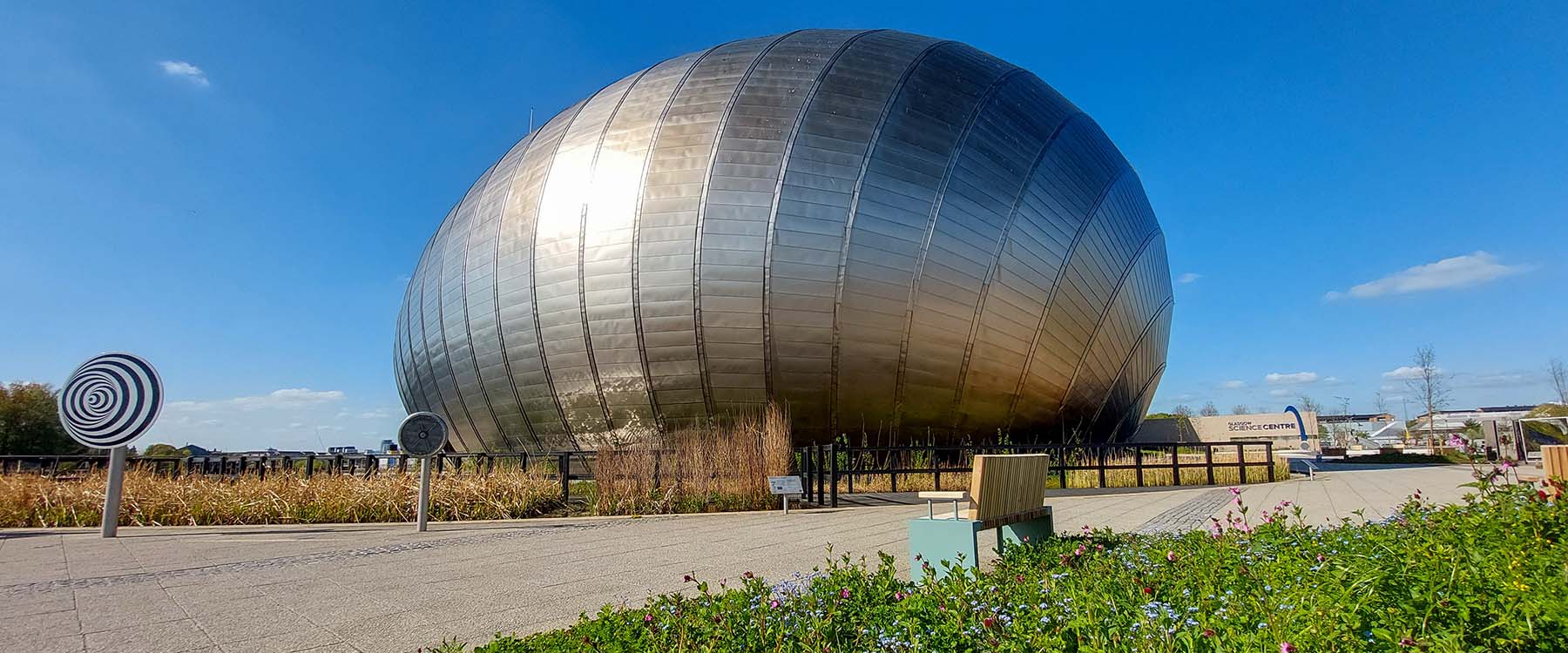 An external view of the IMAX at Glasgow Science Centre on a sunny day. The silver metallic dome is surrounded by a moat and wildflowers. 