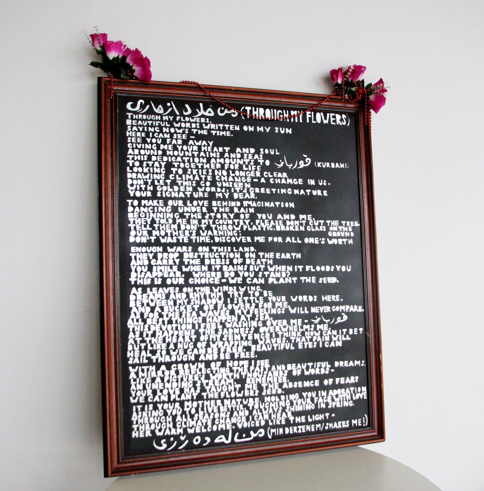 Image of mirror with poem written on it, the frame is wooden and there are red beads and pink flowers at the top.