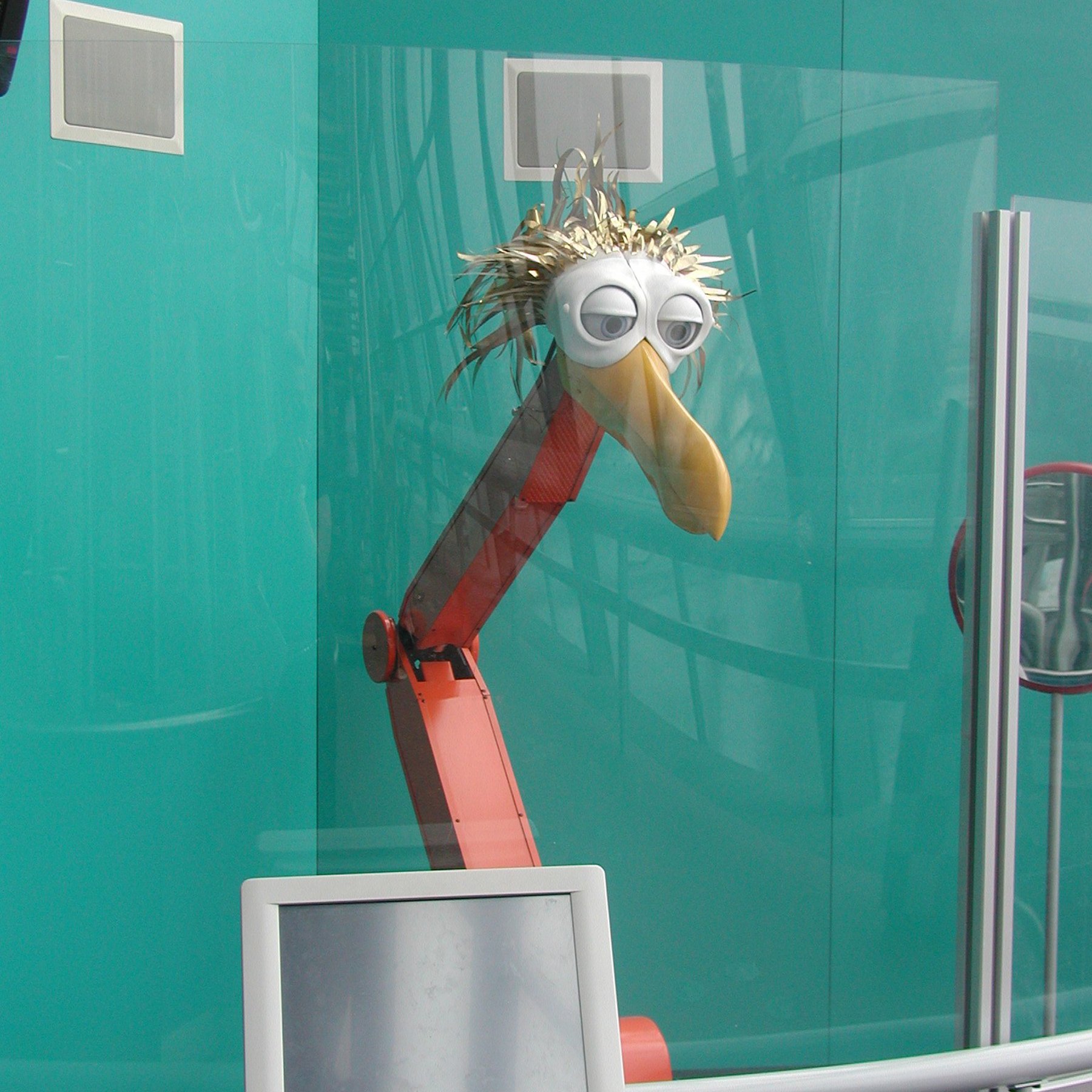 Bird Bot - an orange mechanical beam with the metal head of a bird with a long orange beak and gold tinsel plumage.