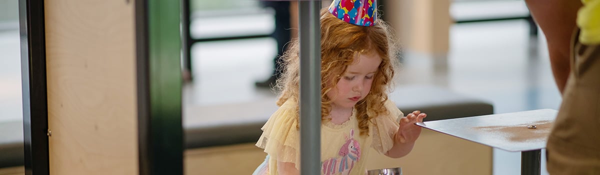 A young girl wearing a party hat looks into a container of sand with great curiosity.