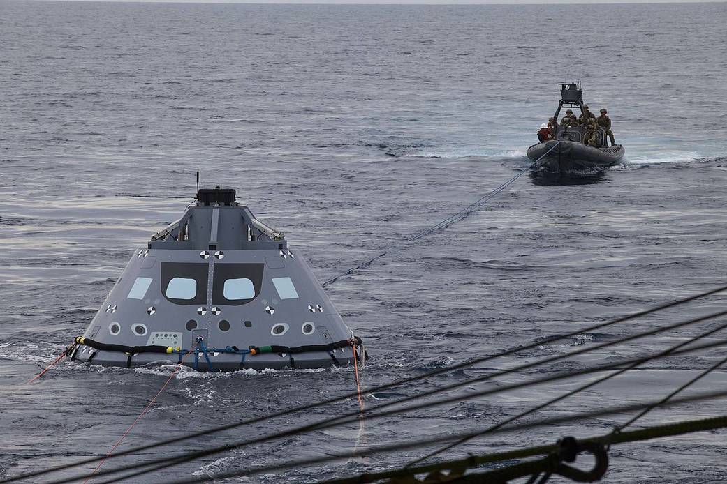 U.S. Navy divers and other personnel in a rigid hull Zodiac boat have attached tether lines to a test version of the Orion crew module during Underway Recovery Test 5 in the Pacific Ocean
