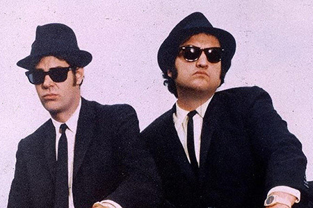 Two men wear sunglasses, black hats and suits.