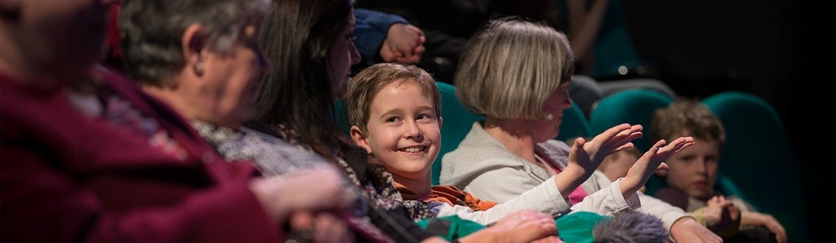 an audience in the science show theatre. A child looks at their parent whilst shrugging their shoulders in amazement