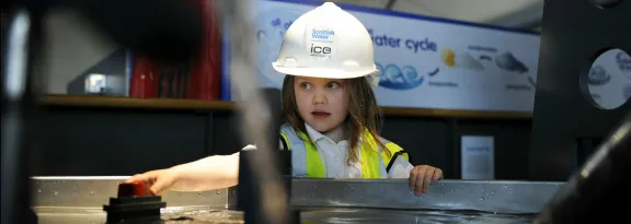 A girl wearing a helmet and high vis vest plays at a water exhibit