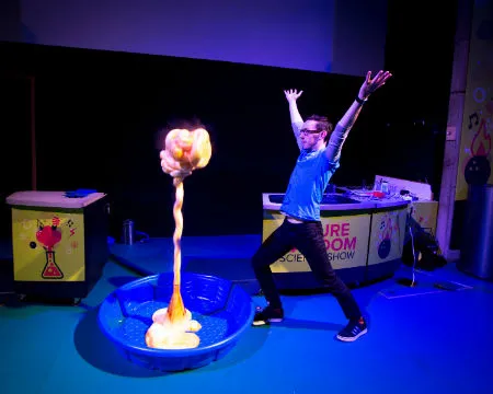 A science communicator on stage performing the Elephant's Tootpaste demo. A plume of 'toothpaste' rises up to almost the same height as the presenter.