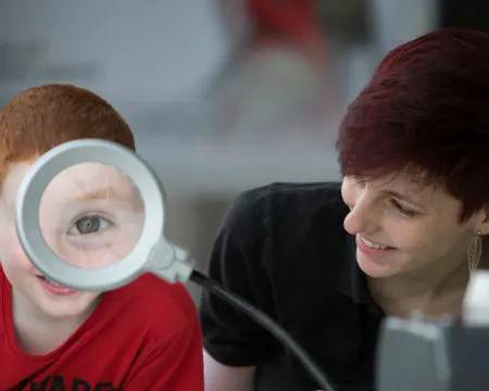 A boy's eye appears huge through a magnifying glass. An expert sits beside him.