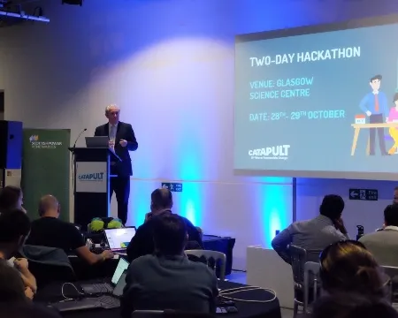 A speaker addresses a seated audience at the hackathon