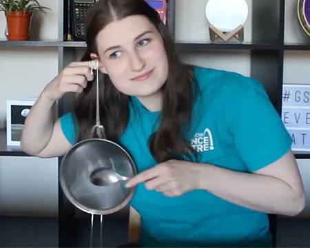 Presenter Natalie holds a piece of string, attached to a kitchen utensil, to her ear