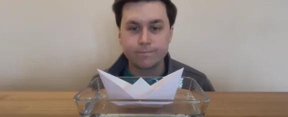 Ross looks at a paper boat floating on water.