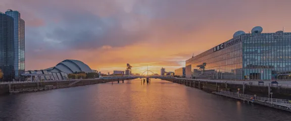 A photo of the sunrise looking east along the River Clyde from a bridge. The sky is yellow, blue and purple and reflected on the surface of the water. Image: Paul Upward and Timberplay Scotland.