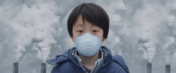 A child wearing a mask stands against a backdrop of plumes of gas rising into the sky from tall chimneys