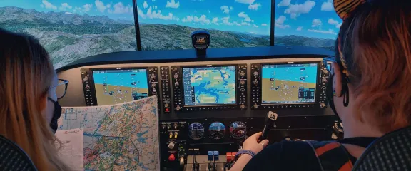 Two people at the controls of a flight simulator. One holds a map whilst the other flys. In front of them is a high resolution view of a mountainous landscape.