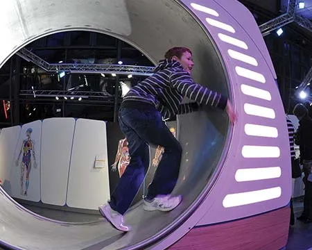 A young person runs in a large metal hamster wheel with lights illuminating a side panel.