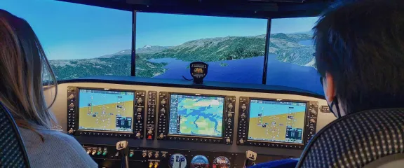 Two people at the controls of a flight simulator look out across a virtual landscape below.