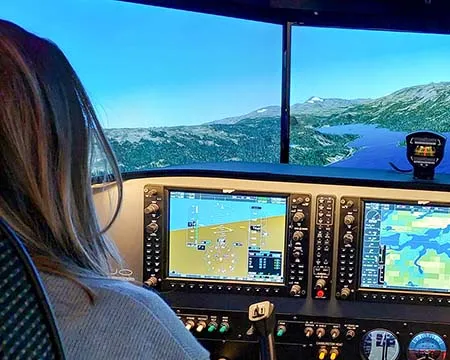 A person at the controls of a flight simulator looks out across a virtual landscape below.