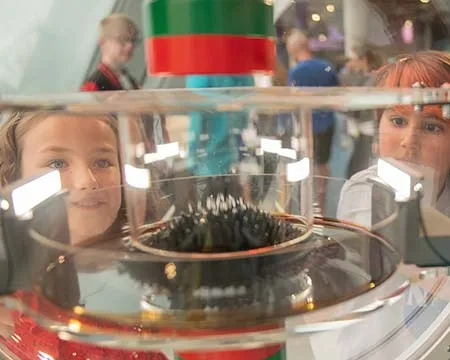 Young people look through to the shape of a magnetic liquid being distorted within a transparent dome