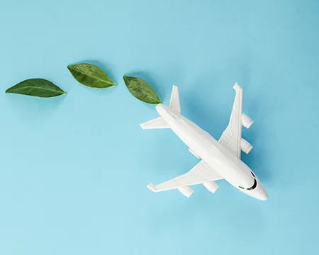 A white paper model of an aeroplane against a blue background. Behind it are a trail of three green leaves.