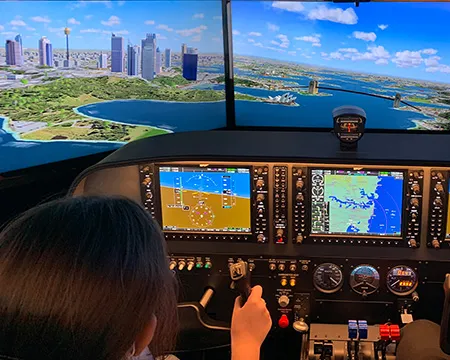 A person at the controls of a flight simulator. In front of them is a high resolution view of a riverside cityscape.
