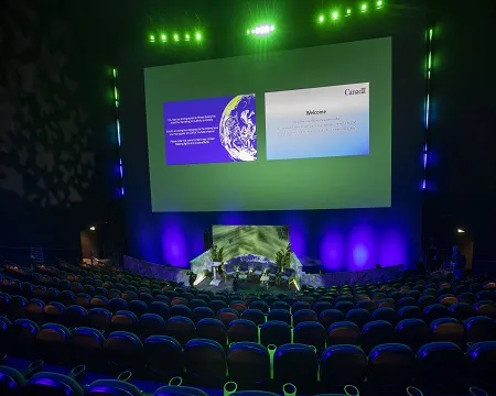 A view over the raked seating of the IMAX theatre towards the giant screen and stage area which are illuminated in purples and greens.
