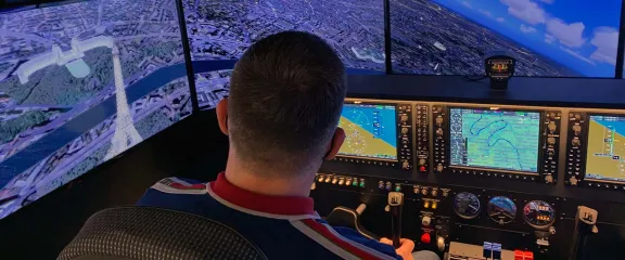 A person at the controls of a flight simulator. In front of them is a high resolution view of a cityscape.