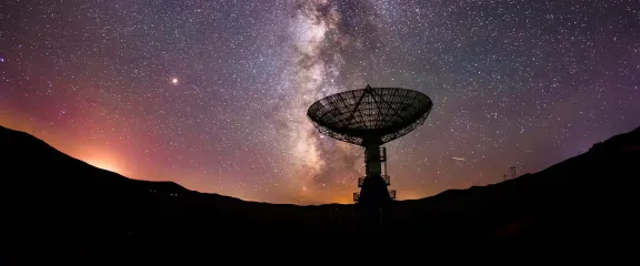 A radio telescope in silhouette against a starry night sky with the Milky Way galaxy centered