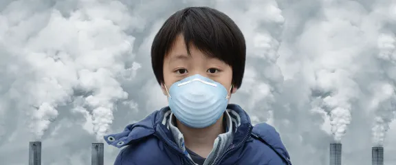 A boy wears a mask. Behind him are grey chimneys with clouds of dust and gas rising into the sky.