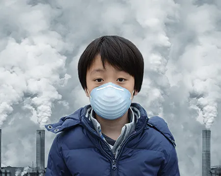 A boy wears a mask. Behind him are grey chimneys with clouds of dust and gas rising into the sky.