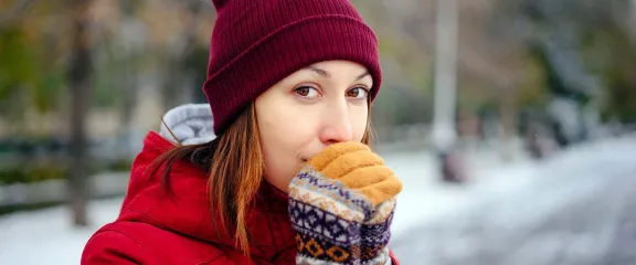 A girl in a red hat and coat, outside in a snowy environment, blows on her gloved hands to warm up her fingers.