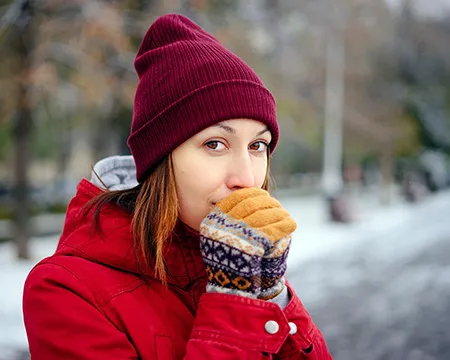 A girl in a red hat and coat, outside in a snowy environment, blows on her gloved hands to warm up her fingers.