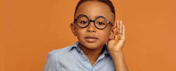 Small boy in glasses holding hand at his ear to listen more closely at something, posing at orange blank wall.