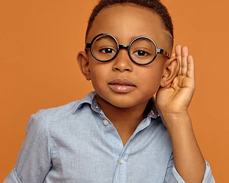 Small boy in glasses holding hand at his ear to listen more closely at something, posing at orange blank wall.
