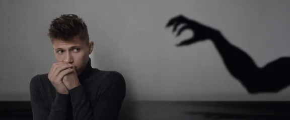 A man with hands clasped and raised to his face looks scared as the shadow of an arm reaches toawards him.