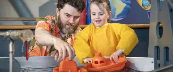 An adult and a child in a yellow apron play with orange toy boats in a water play area, both smiling and engaged.