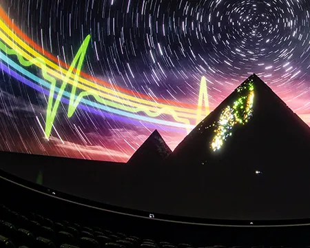 Screen from 'Pink Floyd's Dark Side of the Moon' experience depicting colorful wave patterns and star trails above silhouetted pyramids