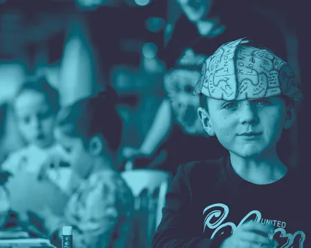 Young boy with a paper brain hat sits in focus, smiling gently. Blurred background shows two children and an adult engaged in activities. 