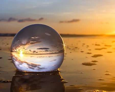 A clear glass ball reflects a vibrant sunset over a beach, capturing clouds and sunlight on a wet, sandy surface, creating a mirrored effect.