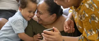 Two female adults with a toddler smile as they look together at a mobile device