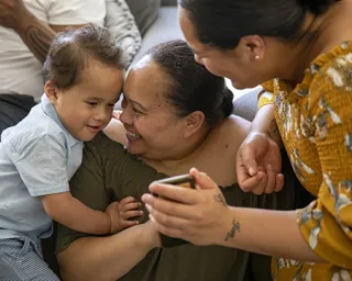 Two female adults with a toddler smile as they look together at a mobile device
