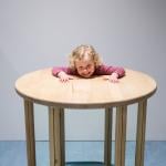A young person with their head and shoulders resting on a wooden table in an illusion that appears to show their 'head on a plate'.