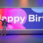 A young person with arms outstretched in front of a giant screen with Happy Birthday and balloons floating across it.