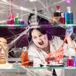 A person in a white lab coat smile from behind a cob-webbed lab containing colourful potions, heads and other Halloween decor.