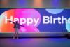 A young person with arms outstretched in front of a giant screen with Happy Birthday and balloons floating across it.