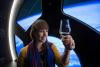 A woman raises a glass of whisky and smiles. They are standing in front of the ISS cupola with a view of Earth in the background.